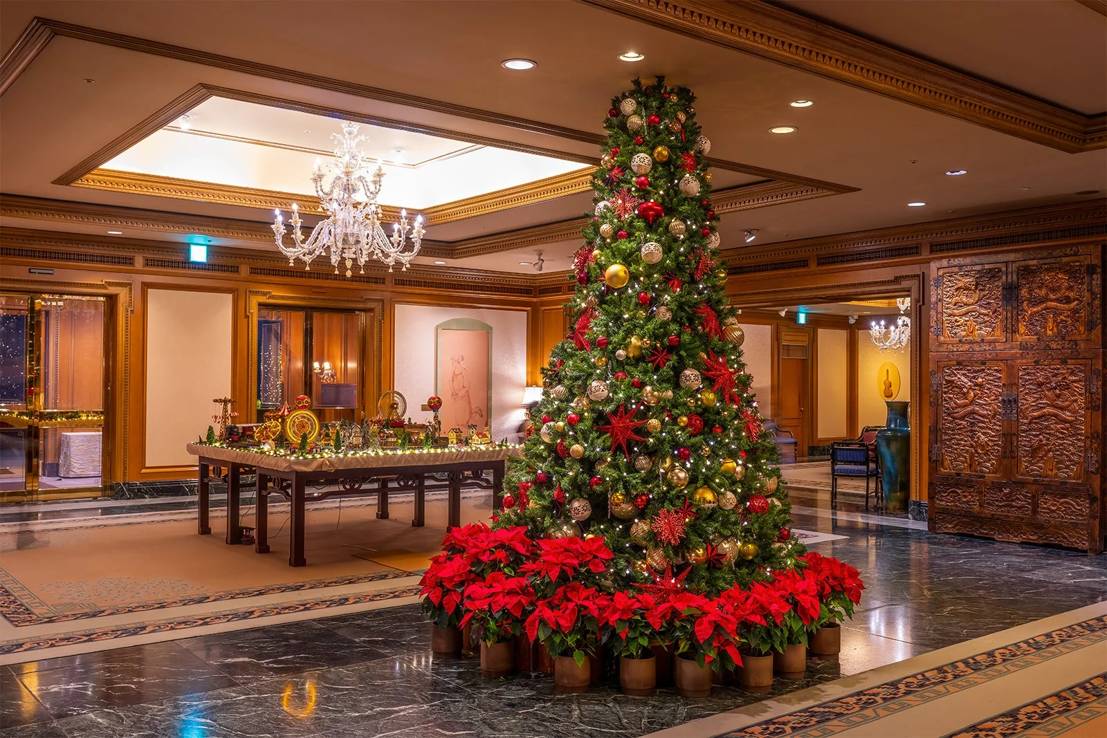 ホテルのロビーに飾られた豪華なクリスマスツリーとポインセチア。金と赤のオーナメントで飾られたツリーが、優雅な空間を華やかに彩っている。天井にはシャンデリアがあり、祝祭的な雰囲気が漂う。