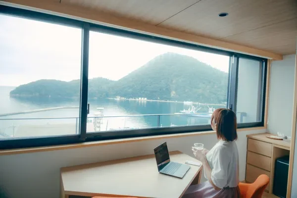 窓から広がる美しい海の景色を眺めながら、女性がノートパソコンに向かっている様子