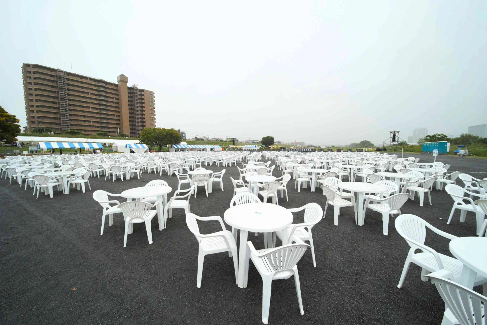 大規模な屋外イベント会場が準備されており、黒い地面に無数の白いプラスチック製テーブルと椅子が整然と並べられています。背景には大きなアパートやストライプのテントが立ち並んでいます。