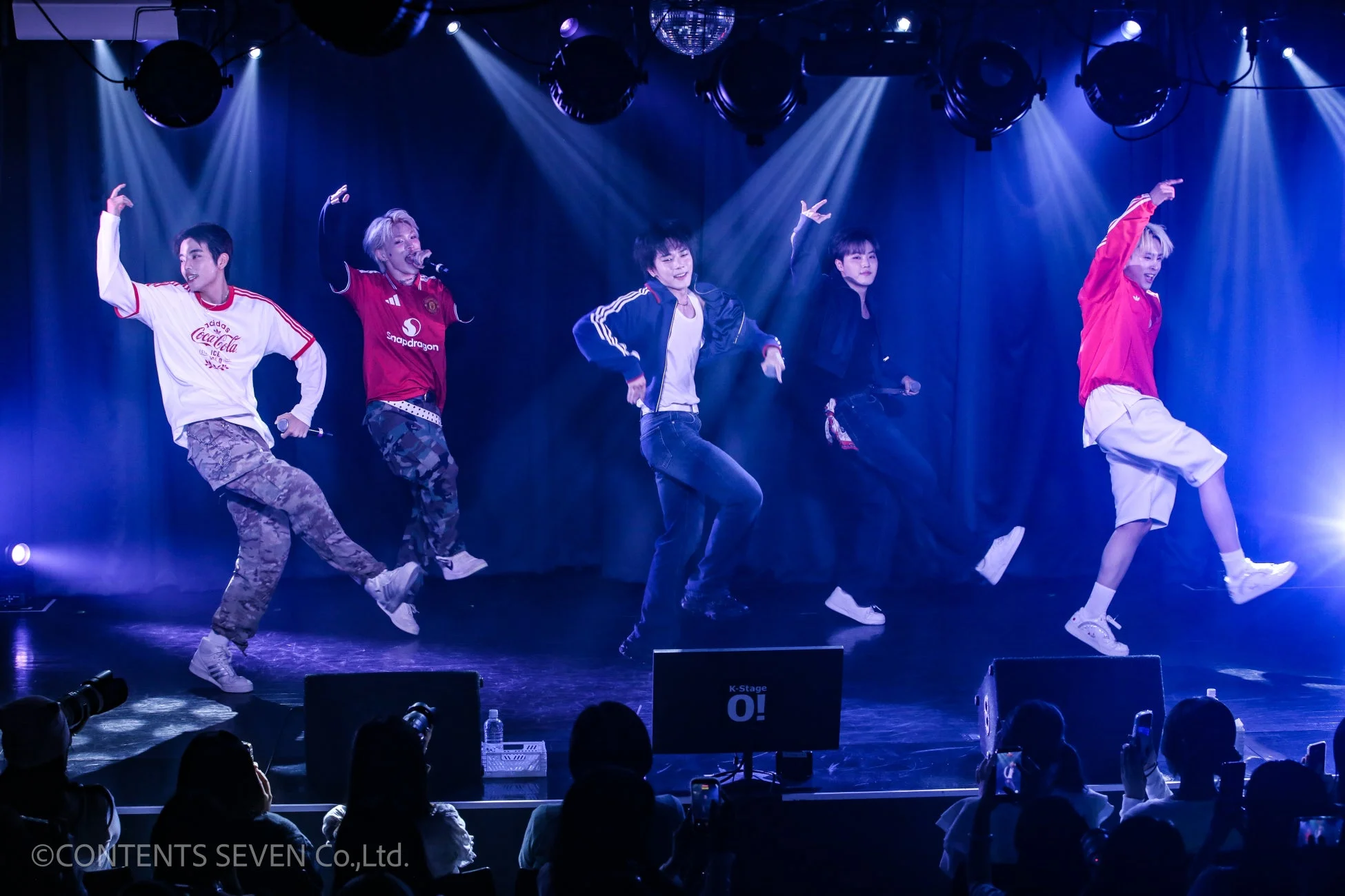 青いスポットライトが照らすステージで、5人の男性パフォーマーがエネルギッシュに歌い踊っているライブパフォーマンスの様子。観客が携帯電話で撮影しており、会場の熱気が伝わる。