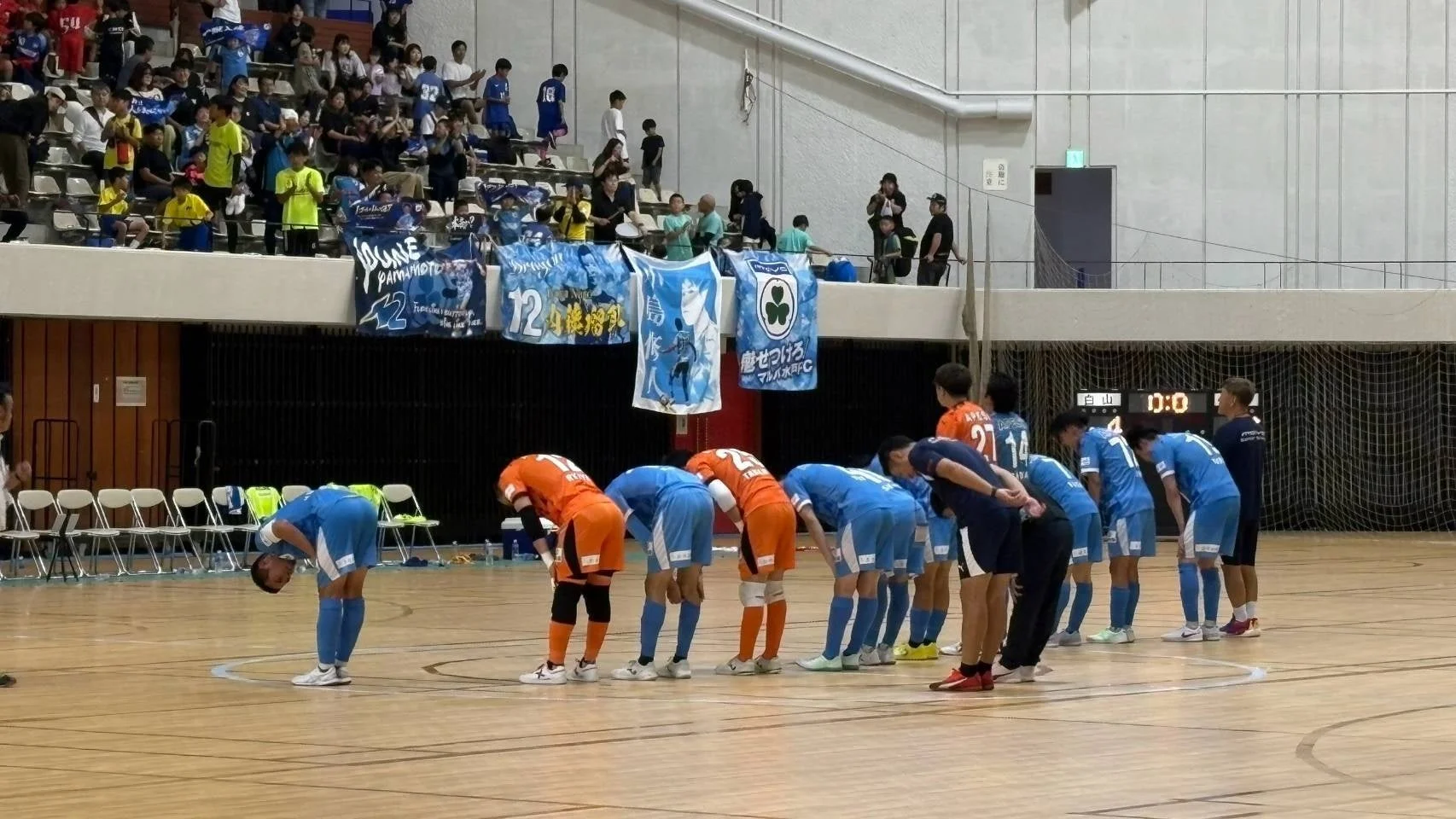 体育館で行われているフットサル試合の様子。選手たちが一列に並んでお辞儀をしており、観客席には多くの人が見守っています