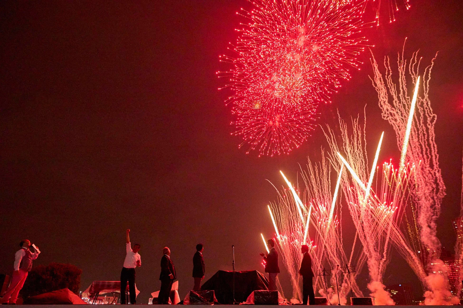 夜空に打ち上げられた赤と白の鮮やかな花火を背景に、ステージ上で数人の人々が立っている。祝賀ムードあふれるイベントの一幕で、一人の人物が拳を突き上げているのが印象的。