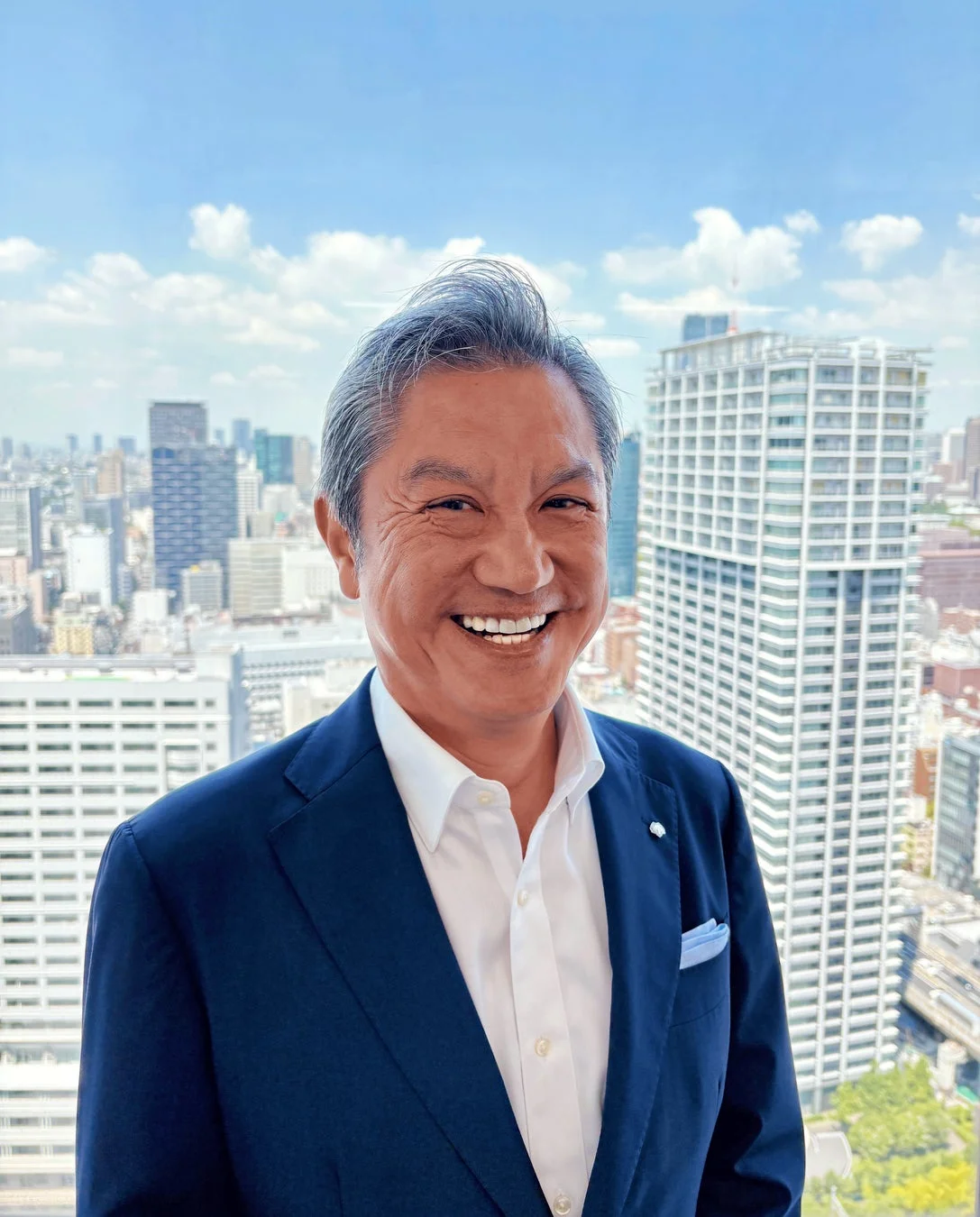 青いスーツを着た笑顔の男性が、高層ビルが立ち並ぶ都市の風景を背景に写っている