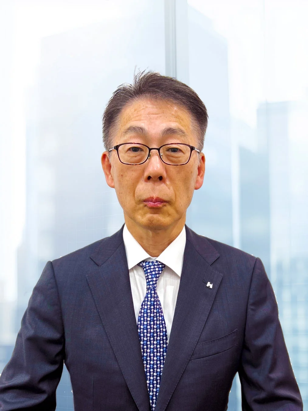 眼鏡をかけた中高年の日本人男性が、紺色のスーツと青いネクタイを着用し、オフィスのような背景で正面を向いて立っている