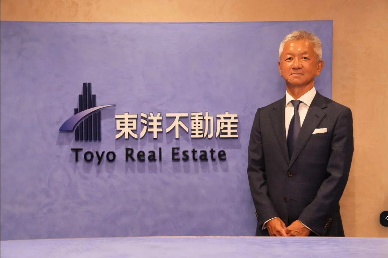 東洋不動産のロゴを背景に、スーツを着用した日本人男性が正面を向いて立っているビジネスシーン