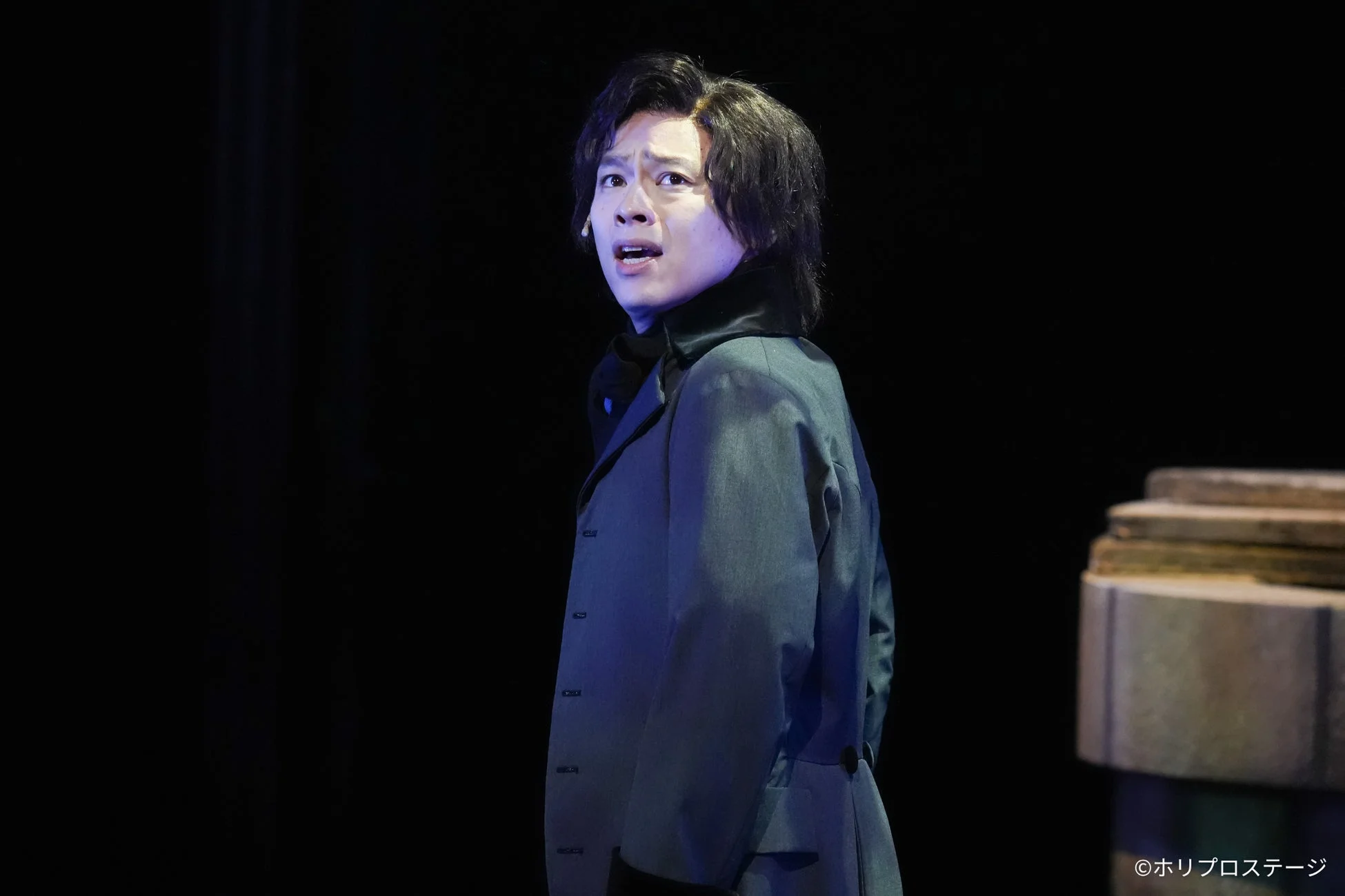 暗い舞台上で、驚きや戸惑いを感じさせる表情で左を向く男性俳優の姿です。黒い襟のコートを着用し、ドラマチックな演技の一場面を捉えています。