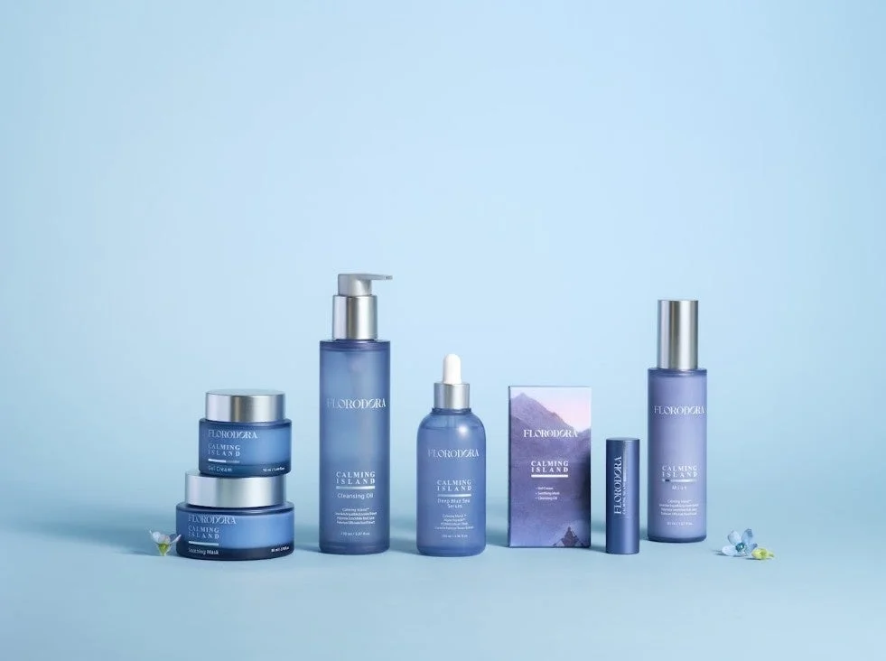 水色の背景にFLODODORA CALMING ISLANDシリーズのスキンケア製品が並べられています。ジェルクリーム、クレンジングオイル、美容液、ミスト、マスクなど、青を基調とした統一感のあるパッケージが特徴です。