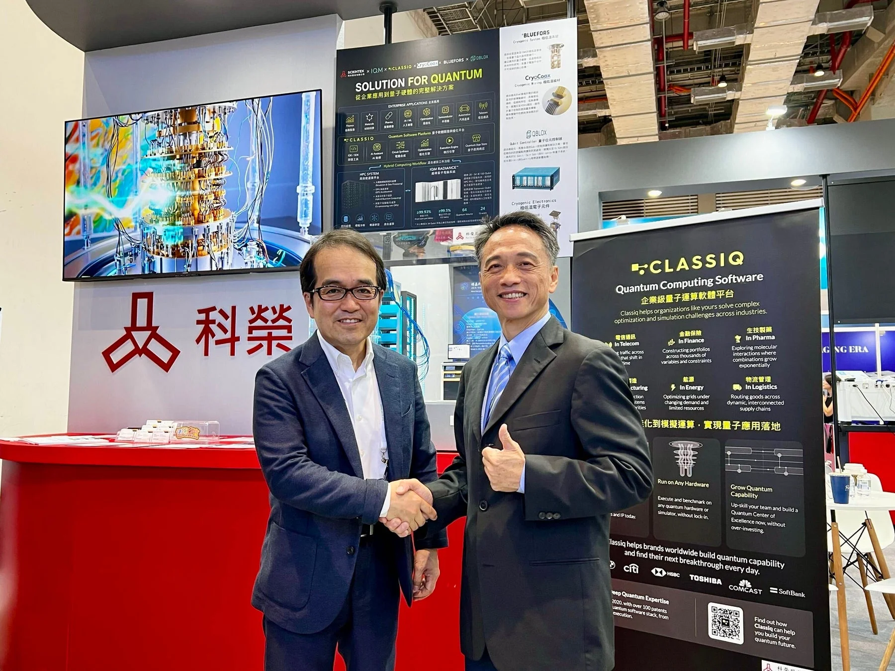 量子コンピューティングの展示ブースで、二人のビジネスマンが握手している様子。Classiqの量子計算ソフトウェアや、様々な企業による量子ソリューションが紹介されており、技術提携やビジネス協業の場面を示しています。